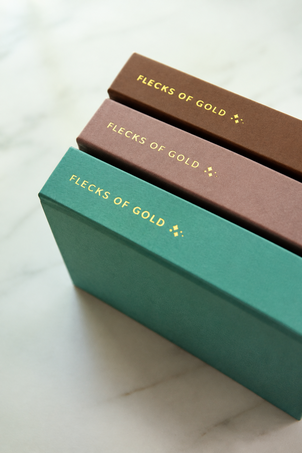 Angled view of camel, rose and teal Flecks of Gold journals stacked to show their gold-foil spines