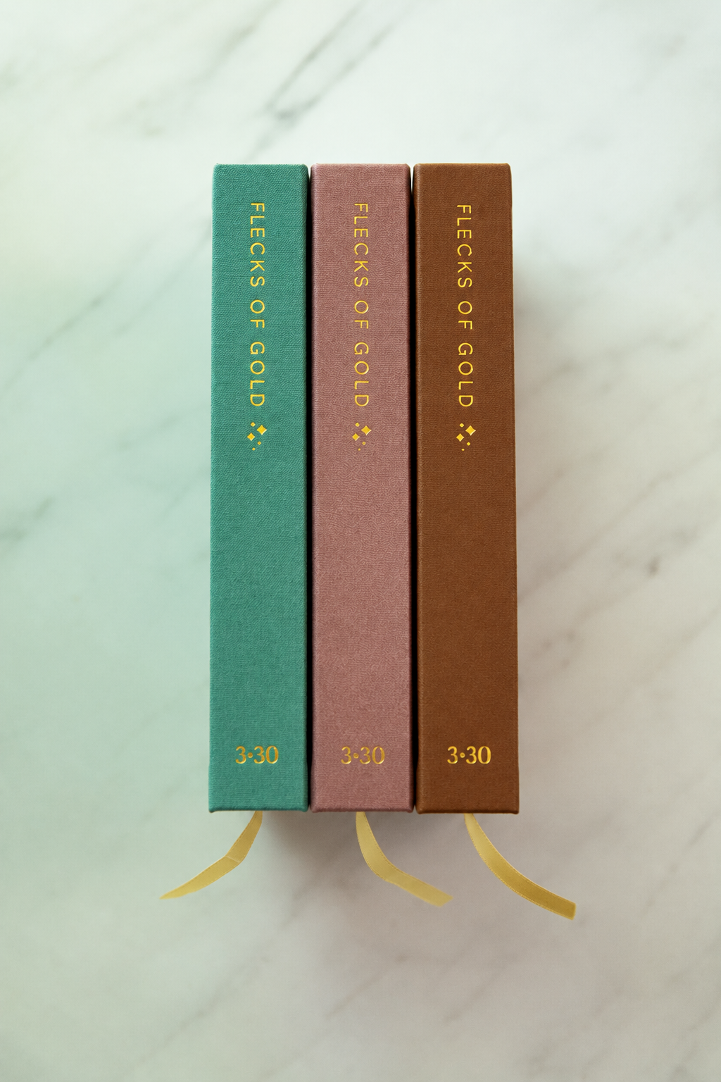 Spines of the teal linen, rose vegan leather and camel vegan leather Flecks of Gold journals standing side by side with gold ribbon bookmarks